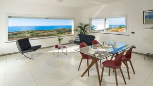 A modern dining area with a glass table, red chairs, and a black sofa. Large windows offer a view of the sea and landscape. - Villa Lily with private pool - My Rental Homes (Montallegro)