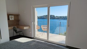 Et soveværelse med seng, skrivebord og stol. Der er en skydedør, der fører ud til en balkon med udsigt over vand og bygninger.