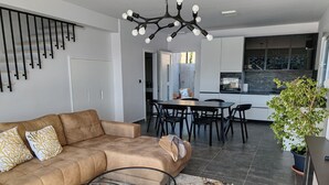 En moderne stue med en beige sofa, et sofabord i glas og et spiseområde med et sort bord og stole. Et køkken er synligt i baggrunden med et stort vindue.