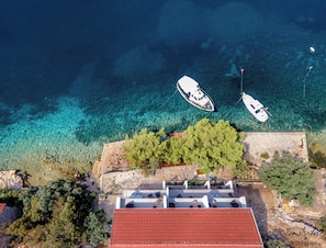 An aerial view of a coastal area with two boats anchored in clear blue waters, a rocky shoreline with trees, and a building with a red roof. - Discover Lastovo (Skrivena Luka)