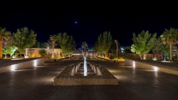 Une vue nocturne d’une place bien éclairée, avec une fontaine centrale, entourée d’arbres et bordée de chemins illuminés.
