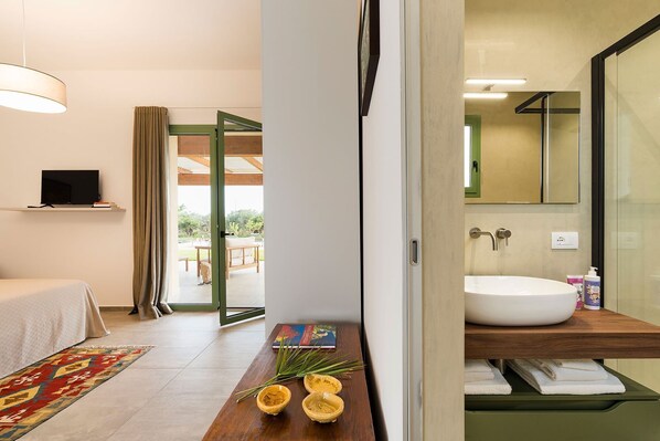 A modern hotel room with a bed, a TV, a wooden side table with books and a plant, a large window, and a bathroom with a sink and mirror.