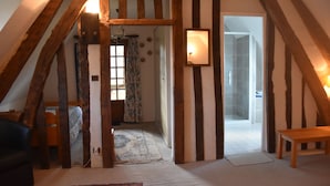 Quarto com vigas de madeira aparente, uma cama, um banquinho e um espelho na parede.