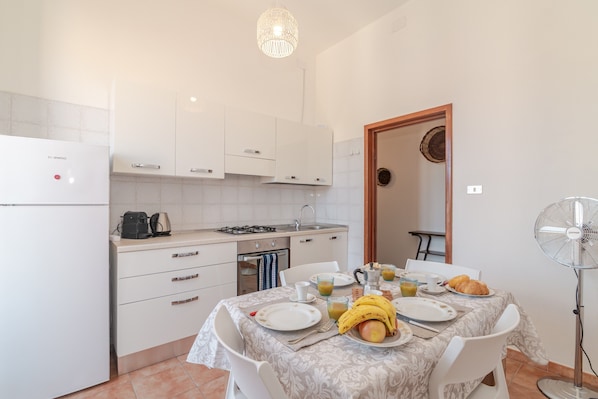 Une cuisine avec des meubles blancs, une table à manger dressée avec de l’assiette, un ventilateur et des aliments pour le petit-déjeuner.