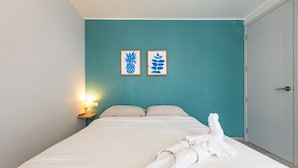 Una cama bien hecha con una toalla blanca, una pared color turquesa y dos cuadros con hojas.