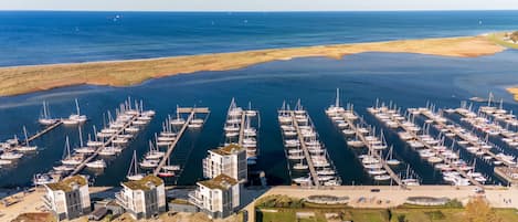 Vista aérea de un puerto deportivo con numerosas embarcaciones amarradas en una disposición simétrica, rodeado por un paisaje costero y un conjunto de edificios con estacionamiento y zonas peatonales.