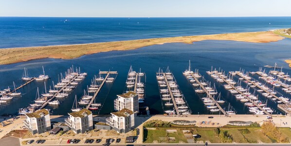 Une vue aérienne d’une marina où de nombreux bateaux sont amarrés selon un plan en réseau, le tout étant entouré d’un paysage côtier et d’un ensemble de bâtiments comprenant des aires de stationnement et des espaces piétonniers.