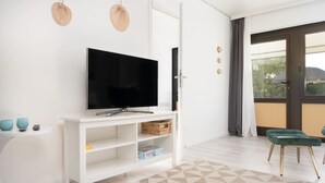 A modern living room with a flat-screen TV on a white stand, a small table with decorative items, a green cushioned chair, and a large window with curtains.