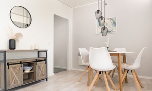 Un coin repas avec une table et des chaises blanches, un meuble en bois sur lequel est posé un bol, un miroir et un luminaire suspendu.