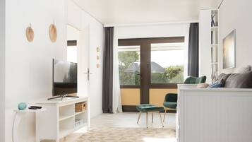 A modern living room with a flat-screen TV, white furniture, a large window with a view, and a patterned floor.