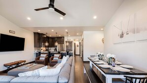 A modern living room with a large grey sofa, a dining area with a long table set for six, and a kitchen in the background. - Family-Friendly Escape w/ Hot Tub & Views | Near Jordanelle & Ski Resorts (Hideout)