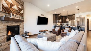 A modern living room with a fireplace, a large sofa, and a kitchen area in the background. - Family-Friendly Escape w/ Hot Tub & Views | Near Jordanelle & Ski Resorts (Hideout)