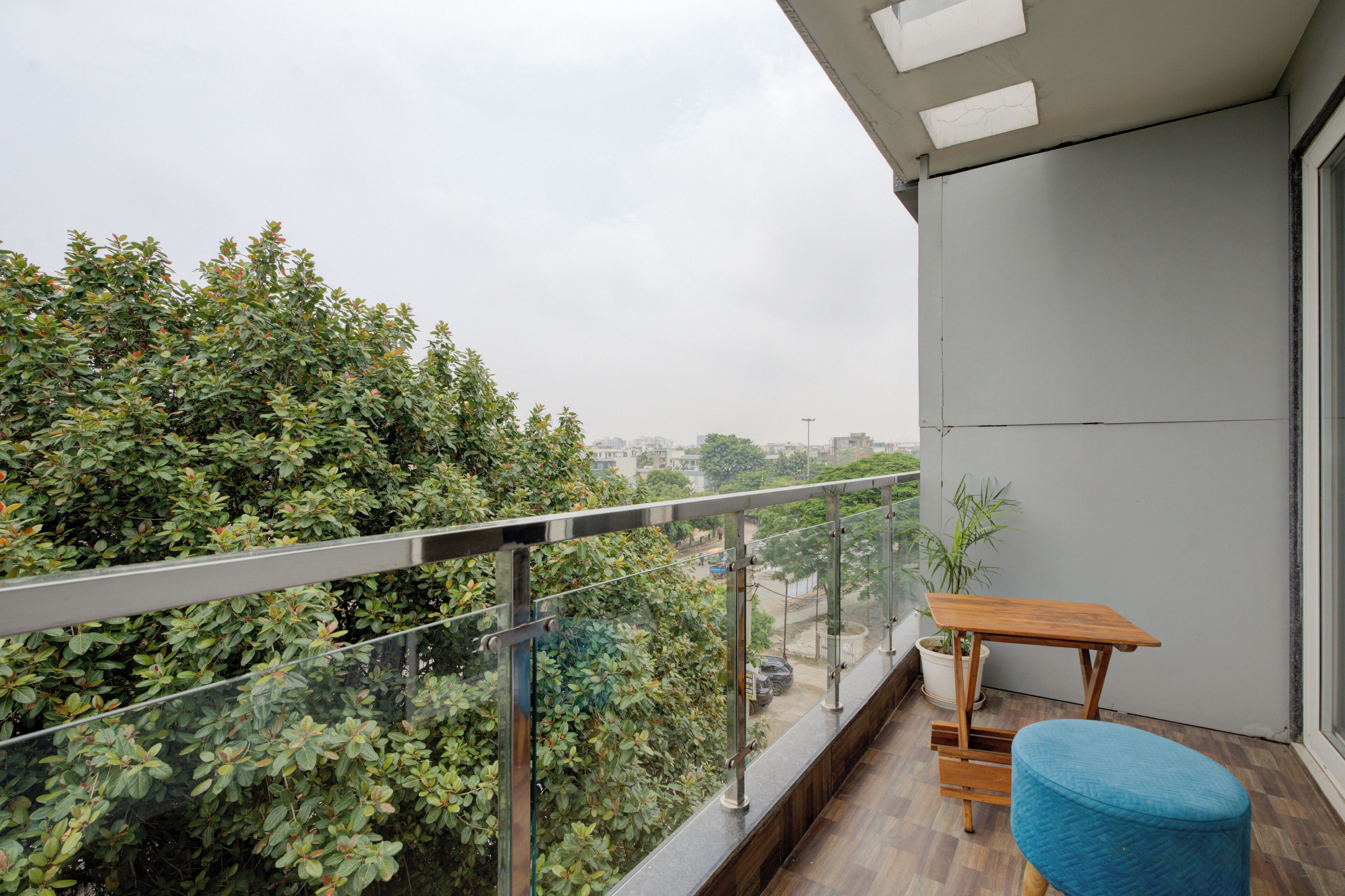 Un balcón con mesa de madera y silla azul, con vista a la vegetación densa y un paisaje urbano.