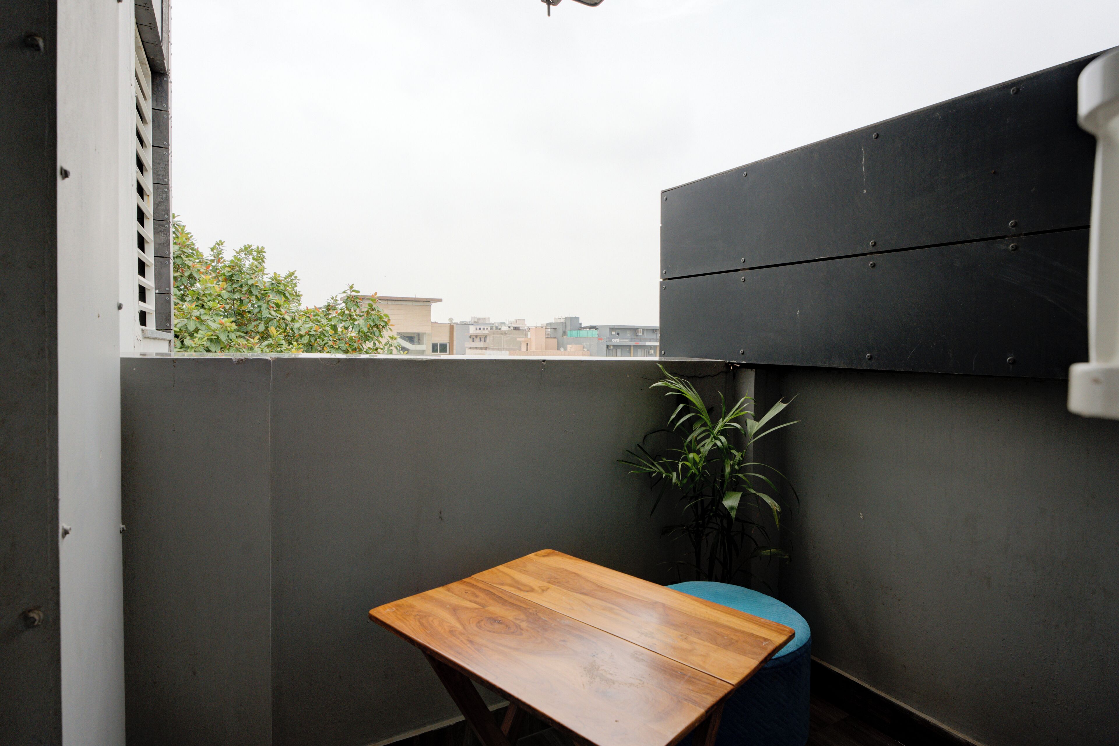 A small balcony with a wooden table, a potted plant, and a view of buildings in the distance.