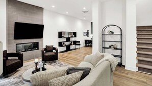 A modern living room with a fireplace, a flat-screen TV mounted on the wall, a coffee table with decorative items, and a staircase with a shoe rack. - Modern Luxury | Fireplace | Rooftop Deck | Near DT (Indianapolis)