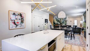 A modern kitchen with a white island, pendant lighting, and a large abstract painting. - Stylish 4BR | Hot Tub | Patio | Firepit | Near DT (Indianapolis)