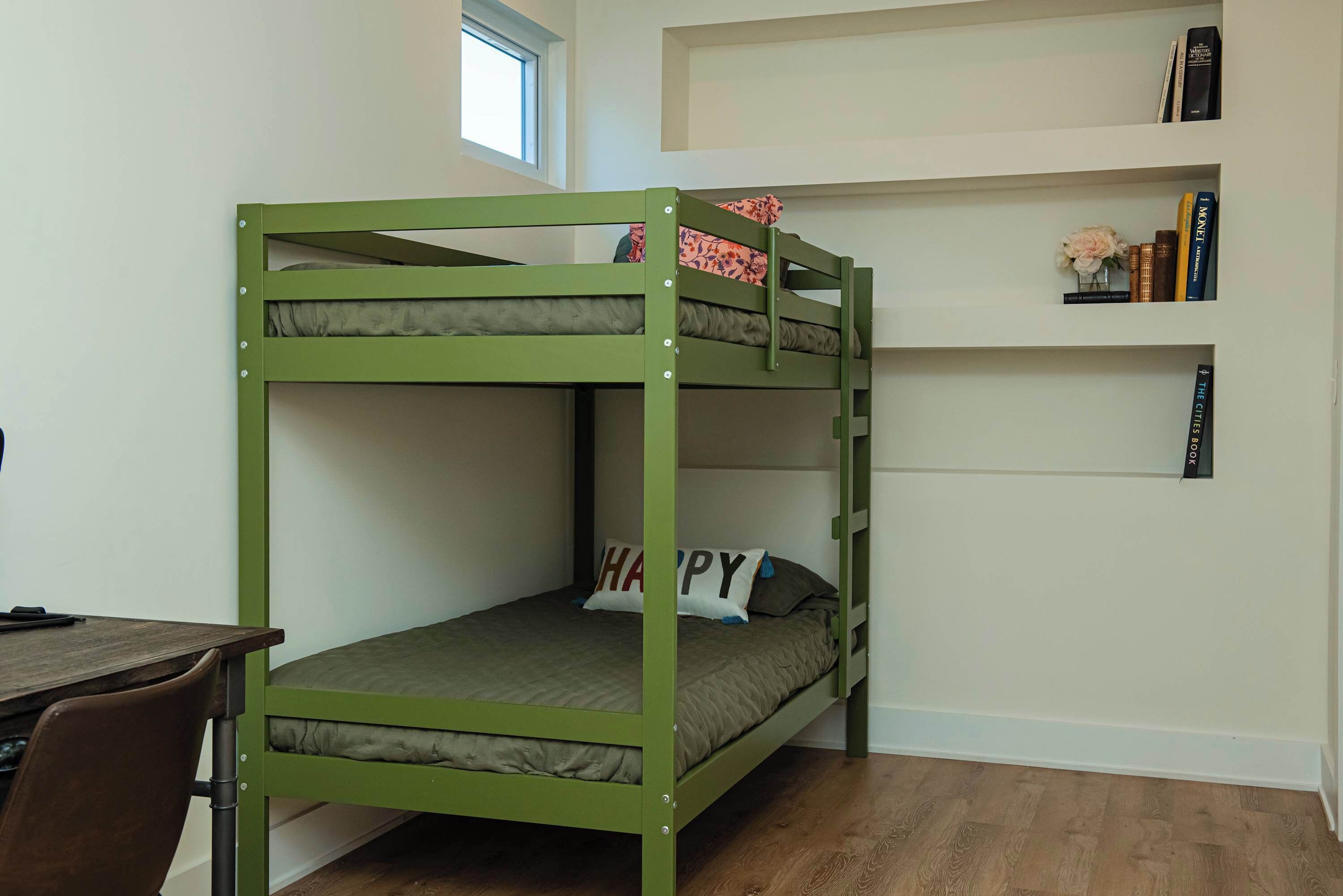A bunk bed with a desk and chair, a bookshelf with books and flowers, and a window in the background.