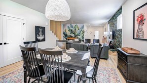 A modern dining area with a dark wooden table and chairs, a hanging light fixture, and a staircase leading to a living area with a colorful mural. - Stylish 4BR | Hot Tub | Patio | Firepit | Near DT (Indianapolis)