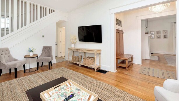 A spacious living room with a staircase, a flat-screen TV, two armchairs, a coffee table, and a wooden console table with baskets.
