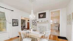 A dining area with a wooden table and white chairs, a chandelier, and a fireplace with framed pictures.