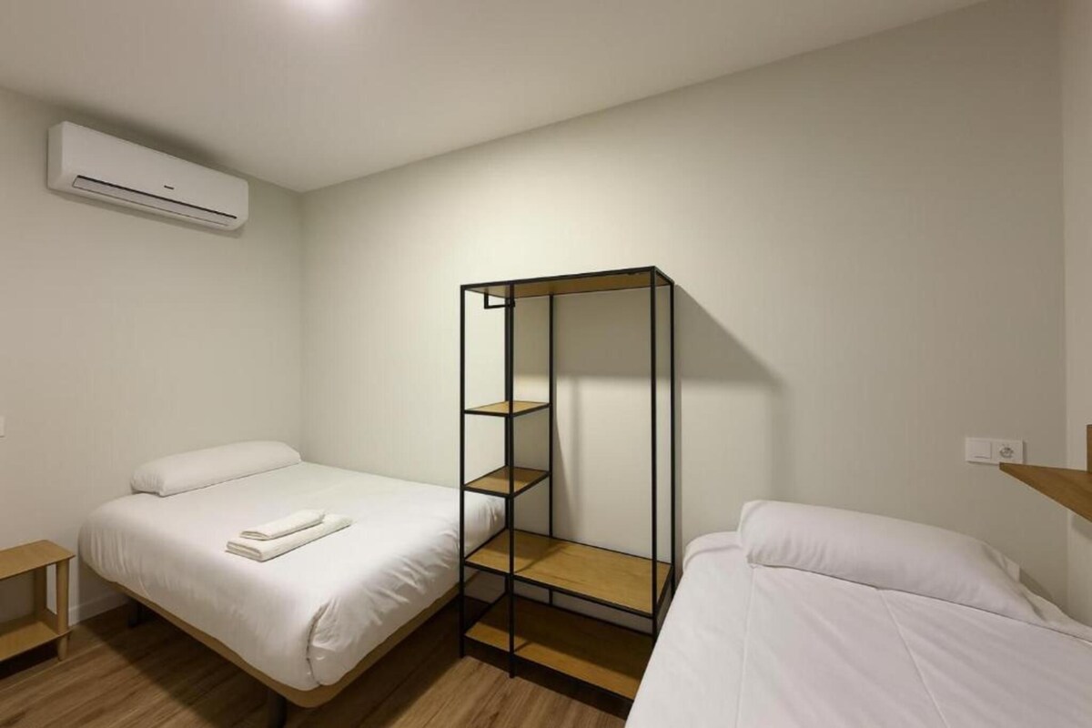 A room with two beds, a wooden floor, a wall-mounted air conditioner, and a metal shelf unit.