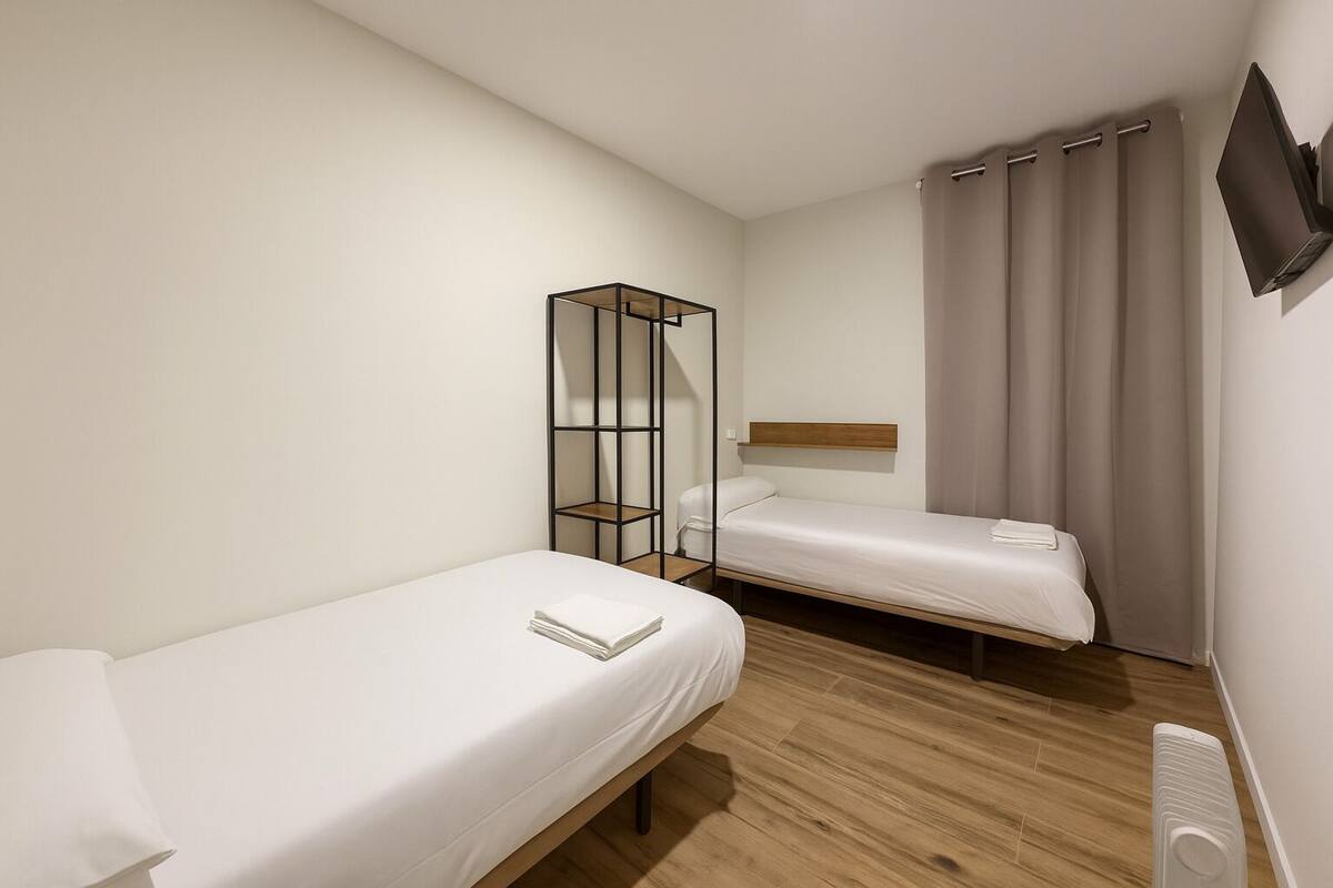 A small, modern hotel room with two single beds, a wooden floor, a built-in TV, and a bedside table.