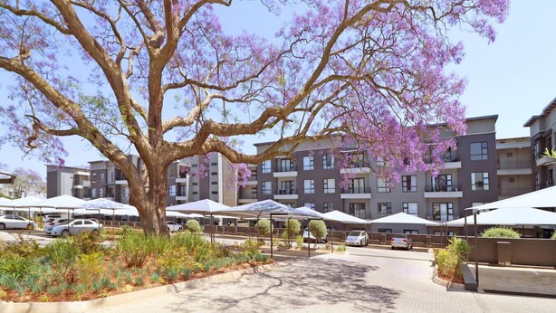 A multi-story residential building with balconies, a parking area, and outdoor seating under umbrellas.