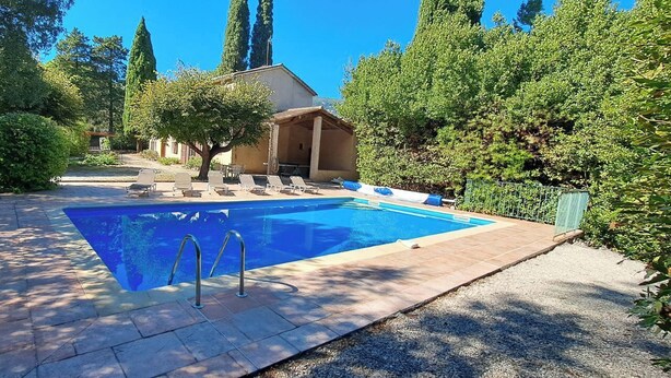 A rectangular swimming pool with a tiled deck, surrounded by trees and a covered seating area with lounge chairs.