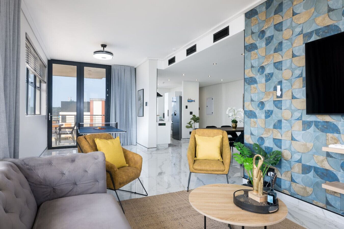 Un salon moderne avec un canapé gris, un fauteuil jaune et une table ronde en bois. Une grande fenêtre offre une vue sur la ville, et un coin repas est aménagé avec une table et des chaises.