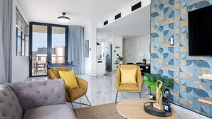 Ein modernes Wohnzimmer mit grauem Sofa, gelbem Sessel und rundem Holztisch. Ein großes Fenster bietet Blick auf die Stadt, und ein Essbereich mit Tisch und Stühlen ist ebenfalls vorhanden.
