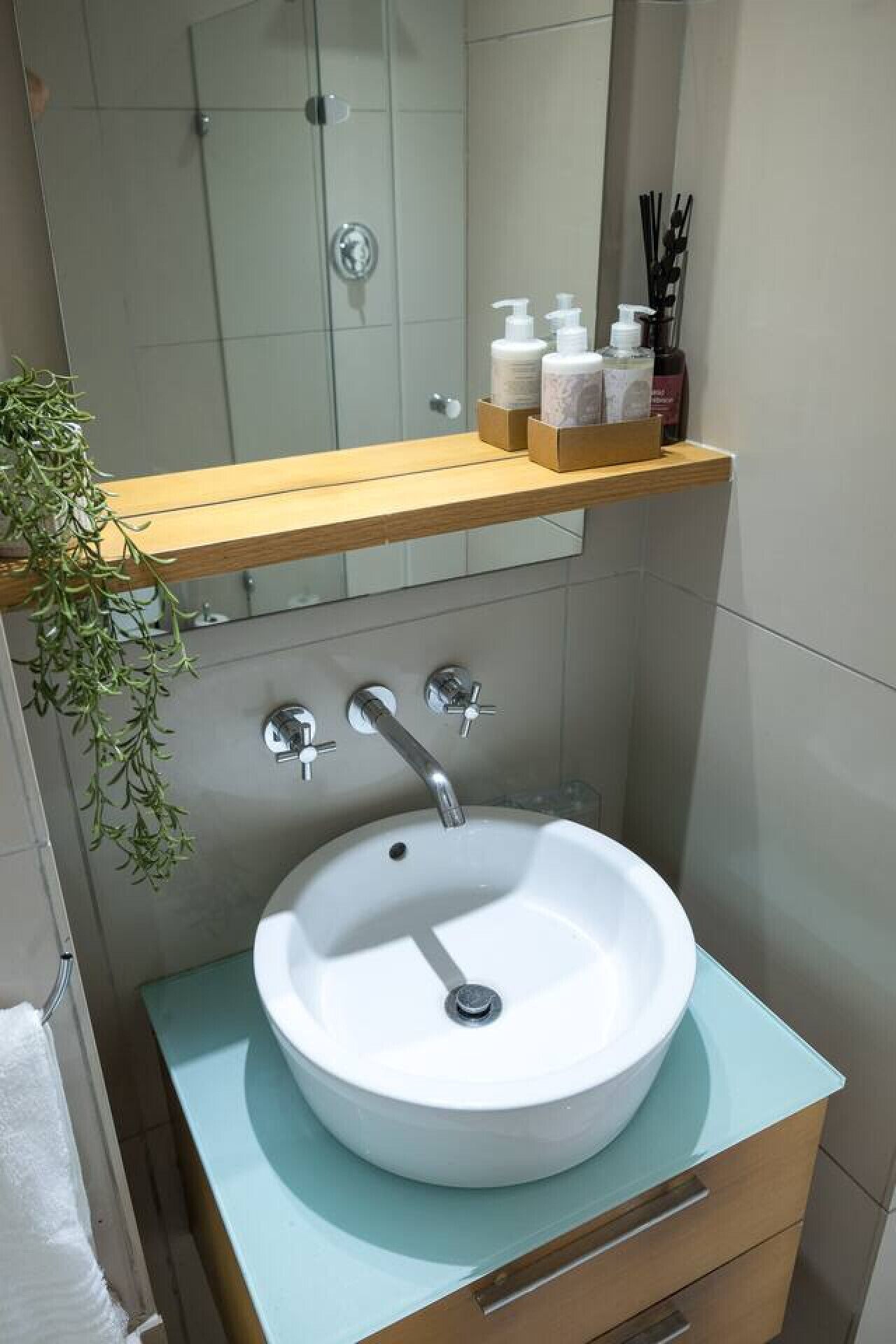 Un baño con lavamanos blanco, dos llaves, un estante de madera con artículos de aseo y una planta colgante.