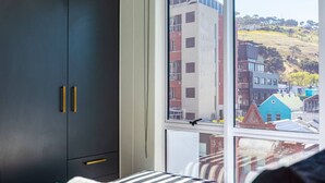 A bedroom with a large window, a bed with striped bedding, and a dark wardrobe with gold handles. - 602 The Valour Luxurious Apartment (Cape Town)