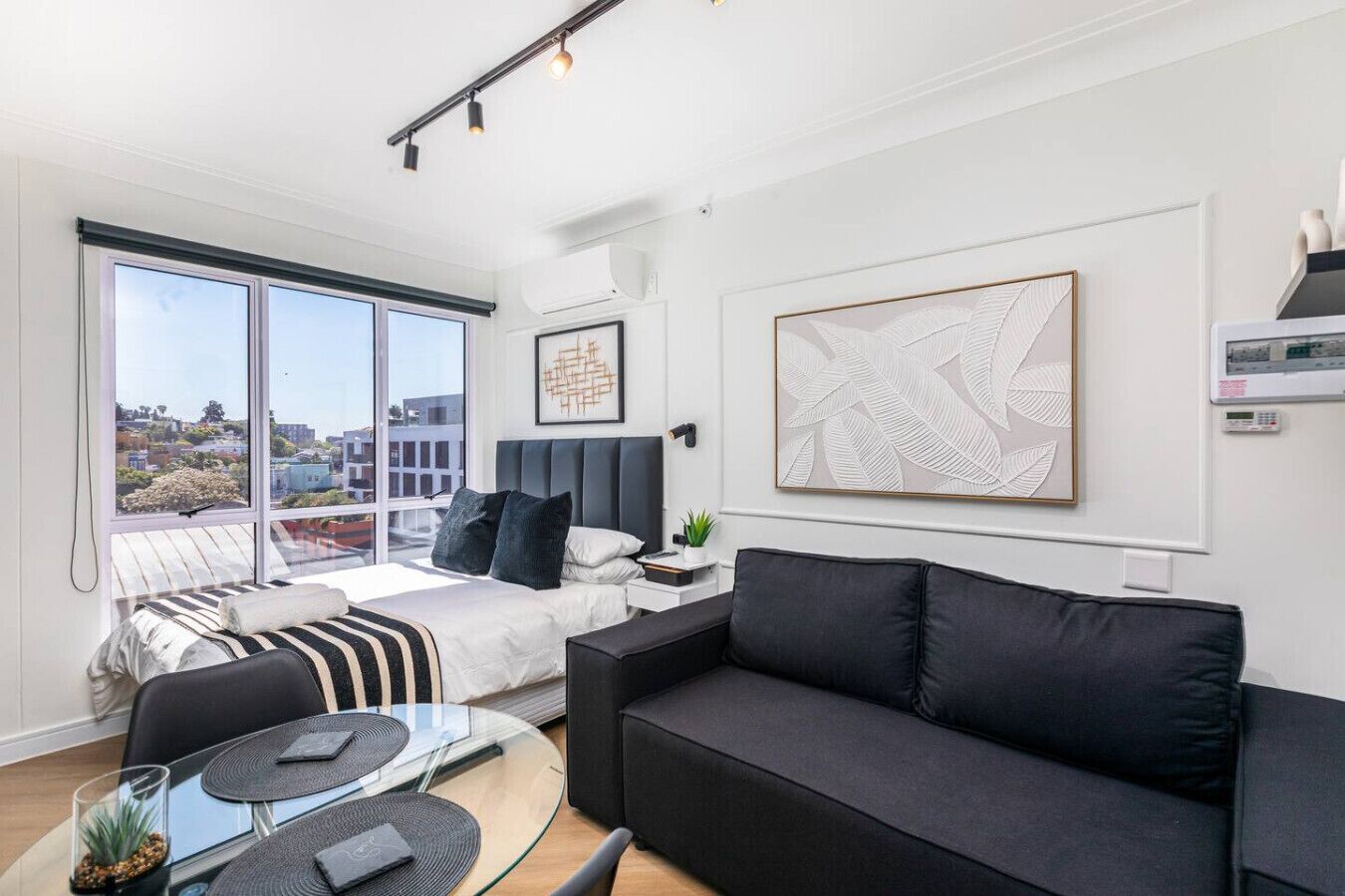 A modern bedroom with a large bed, a black sofa, a glass coffee table, and a city view through large windows.