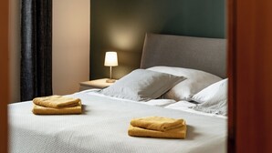 Una cama bien hecha con dos toallas dobladas, una lámpara de noche y una pared de color oscuro.
