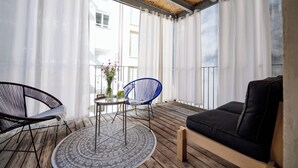 Un balcón con piso de madera, un sofá negro, una silla azul, una mesa redonda y cortinas blancas.