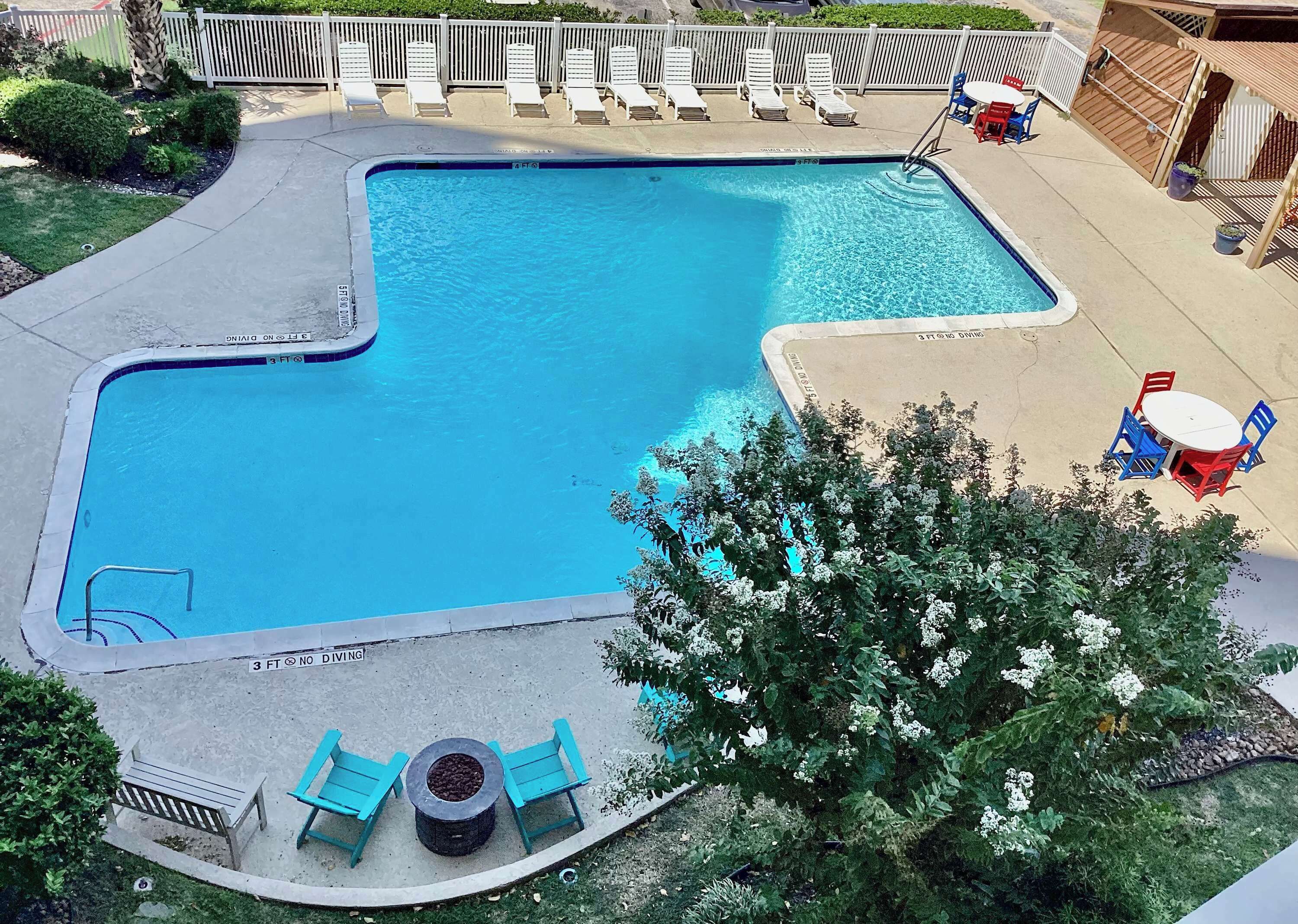 A rectangular swimming pool with a deck area, surrounded by lounge chairs and a small table with chairs.