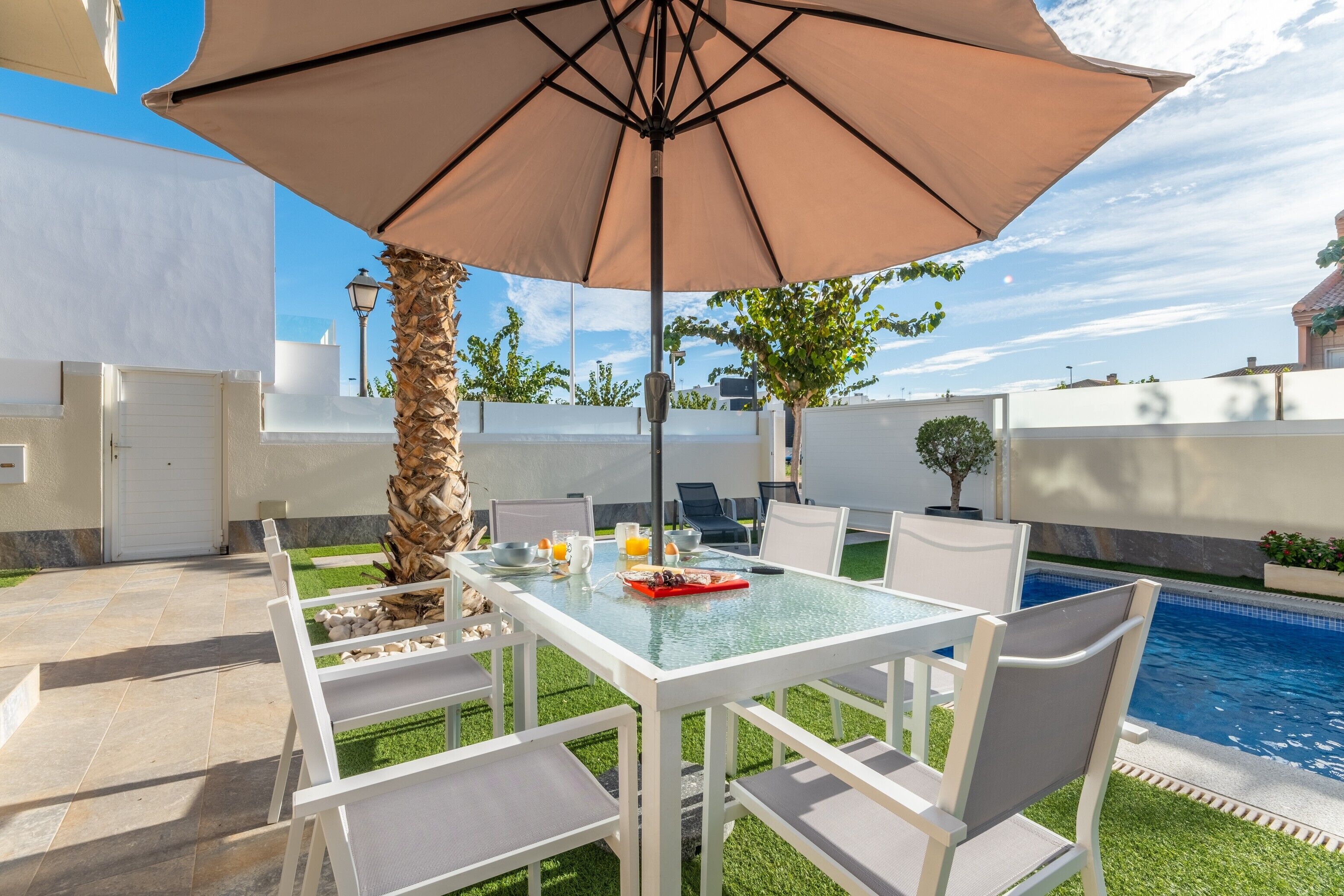 Un patio al aire libre con una mesa de vidrio, sillas blancas, un paraguas grande y una piscina al fondo.