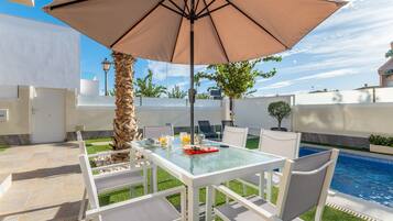 Un patio al aire libre con una mesa de vidrio, sillas blancas, un paraguas grande y una piscina al fondo.