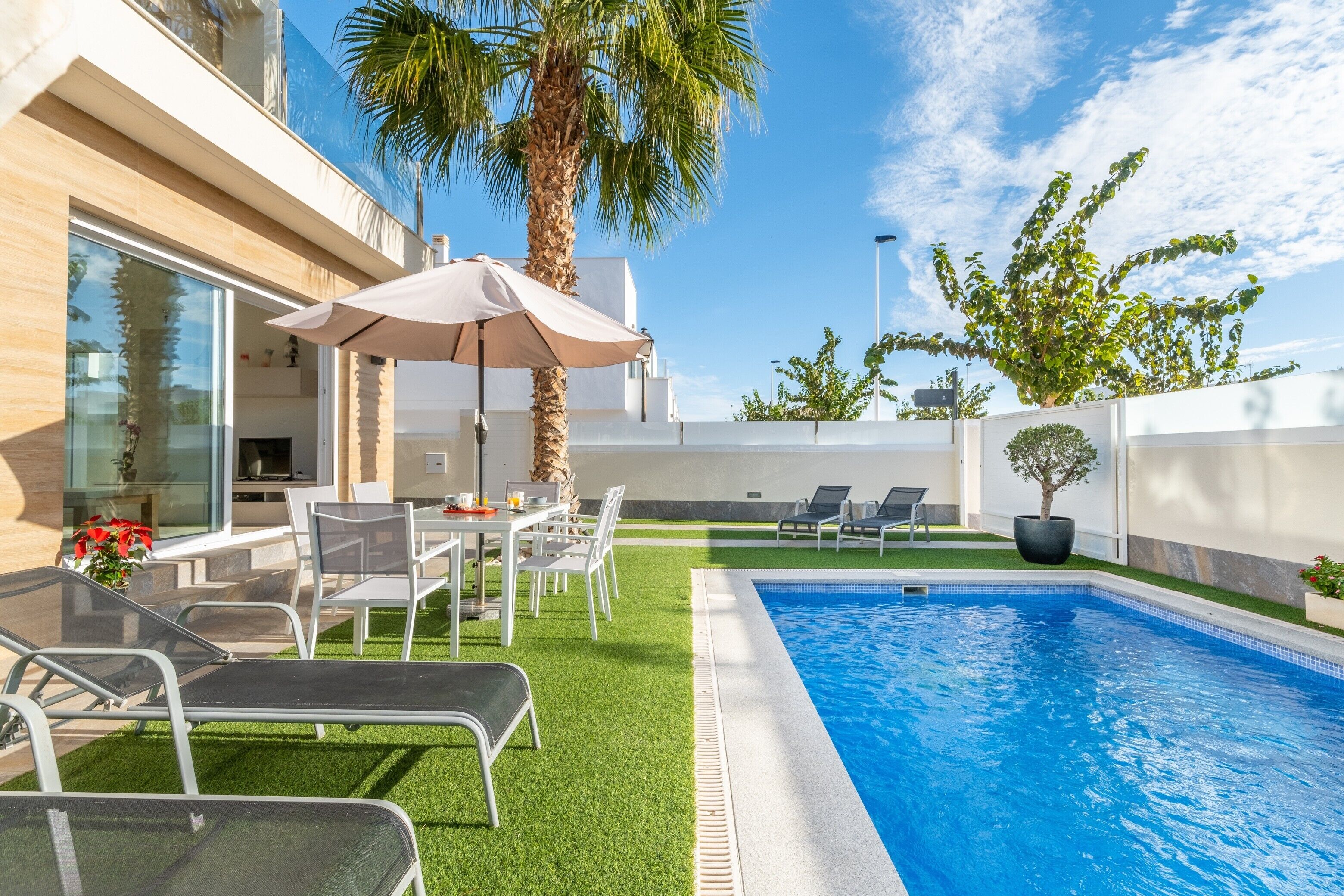 A modern outdoor pool area with a dining setup, lounge chairs, and a palm tree.