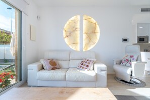 Un salon moderne avec un canapé blanc, une table basse ronde et un fauteuil blanc. Une grande fenêtre se trouve sur un des murs, et on aperçoit une cuisine à travers une porte ouverte.