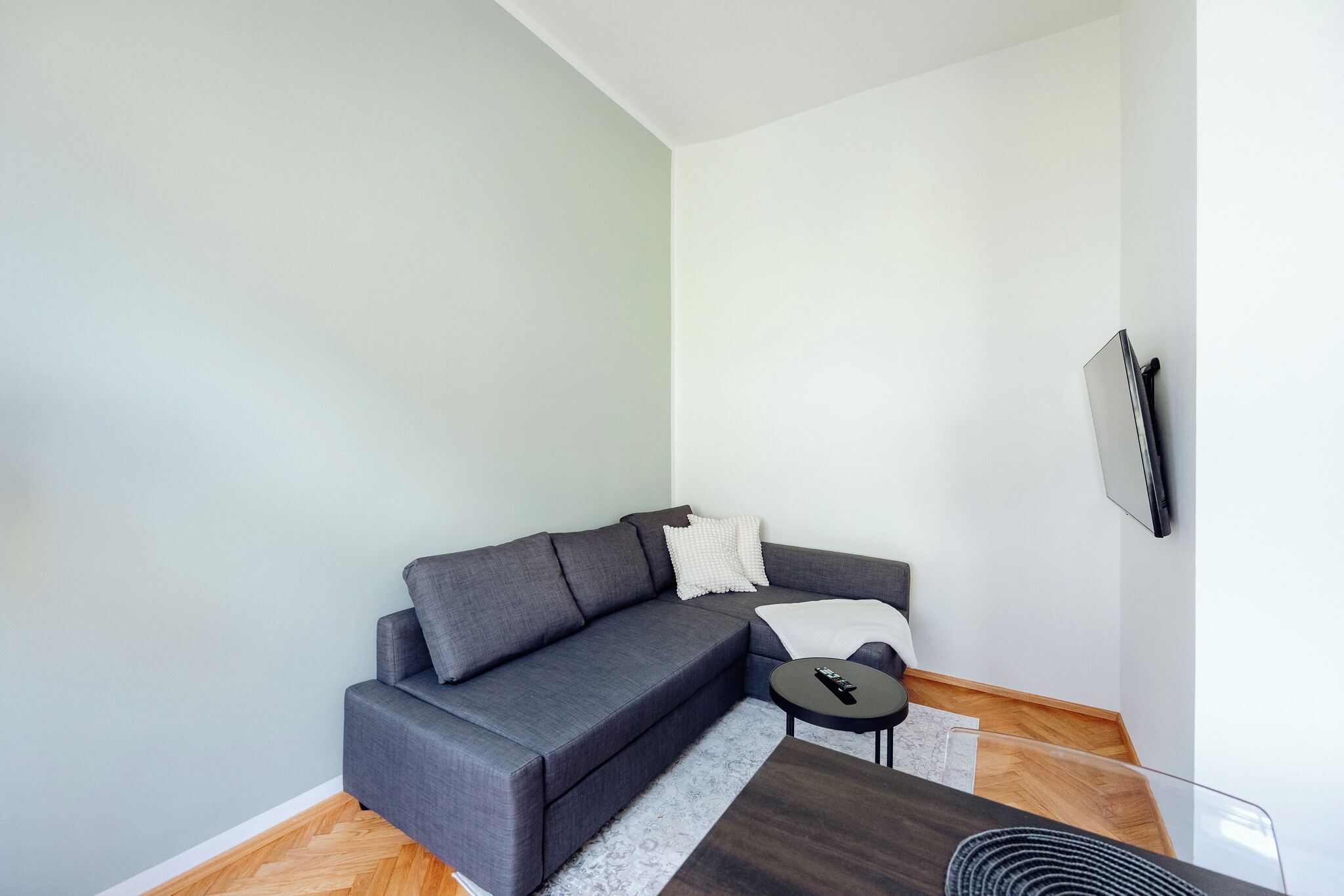 A modern living room with a grey sectional sofa, a small round coffee table, and a flat-screen TV mounted on the wall.