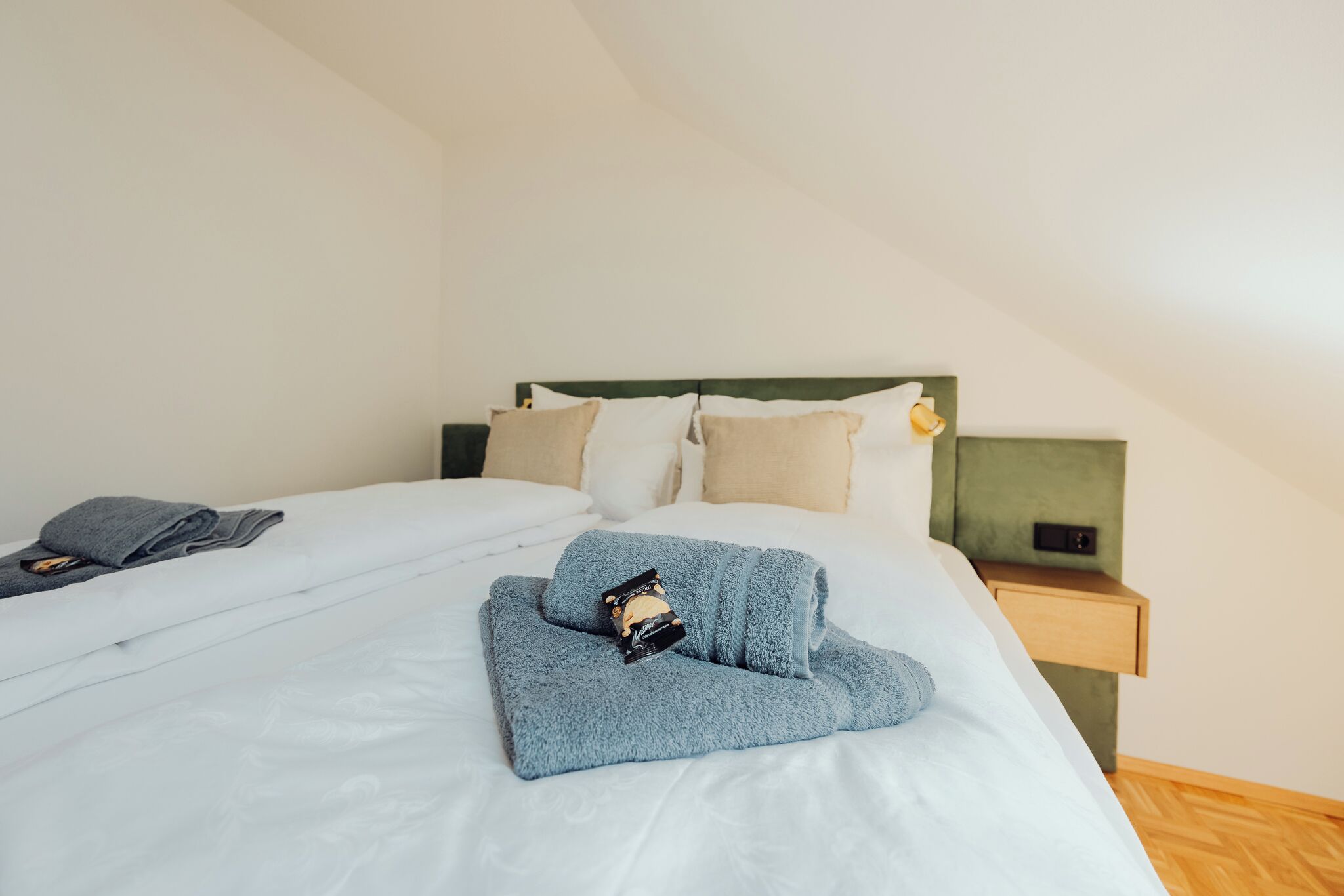 Ein ordentlich bezogenes Bett mit gefaltetem Handtuch und einem kleinen Gegenstand darauf, ein Nachttisch mit Lampe und Holzboden.