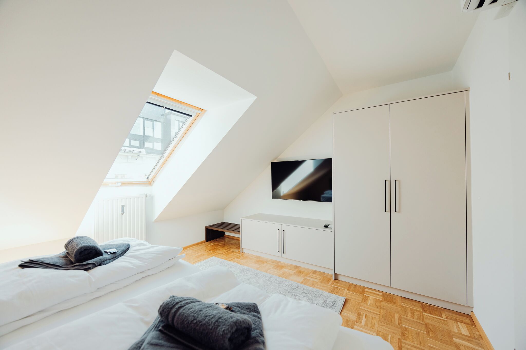 A modern bedroom with a sloped ceiling, a large window, a flat-screen TV, and a white wardrobe.