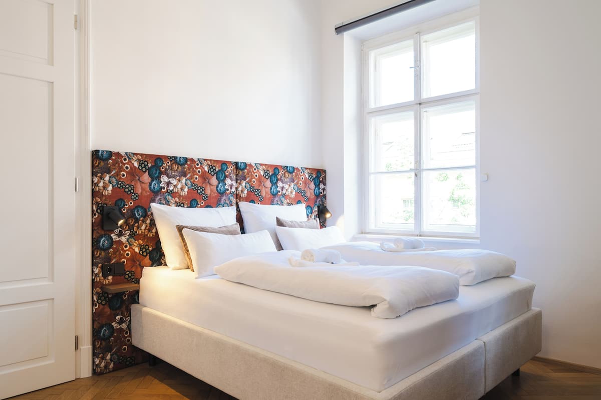 Un lit bien fait, avec du linge de lit blanc et une tête de lit à motifs floraux, dans une pièce lumineuse dotée d’une grande fenêtre.
