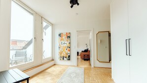 Quarto espaçoso com amplas janelas, piso de madeira em padrão herringbone, tapete cinza, mesa preta e uma pintura abstrata colorida.