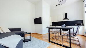 Uma sala de estar moderna com um sofá cinza, uma mesa redonda pequena e um tapete cinza. Há uma área de cozinha com bancada preta, armários brancos e um exaustor preto.