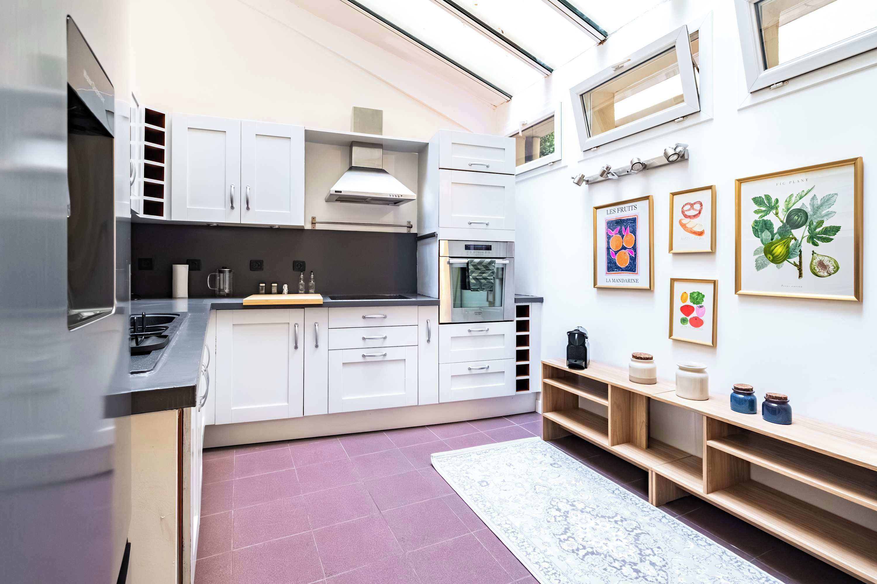 Una cocina moderna con armarios blancos, electrodomésticos de acero inoxidable y encimera de madera. En la pared hay láminas enmarcadas con motivos botánicos y una pequeña estantería con frascos.