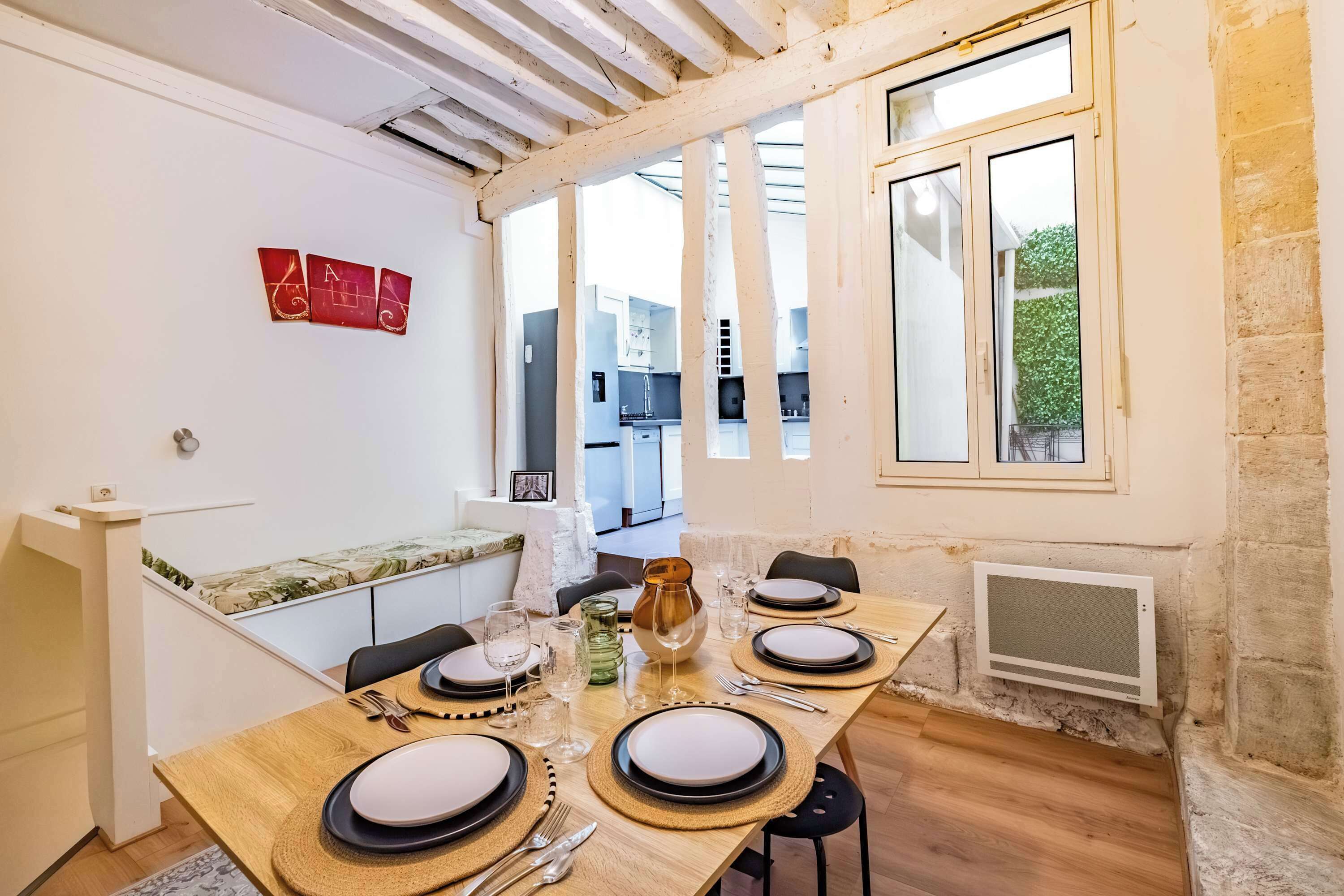Zona pranzo con un tavolo lungo apparecchiato per quattro persone, una cucina sullo sfondo e un'ampia finestra con vista sul verde.