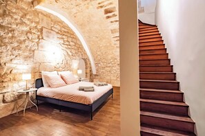 Quarto com parede de pedra, cama com cabeceira, mesa de cabeceira e escada de madeira.