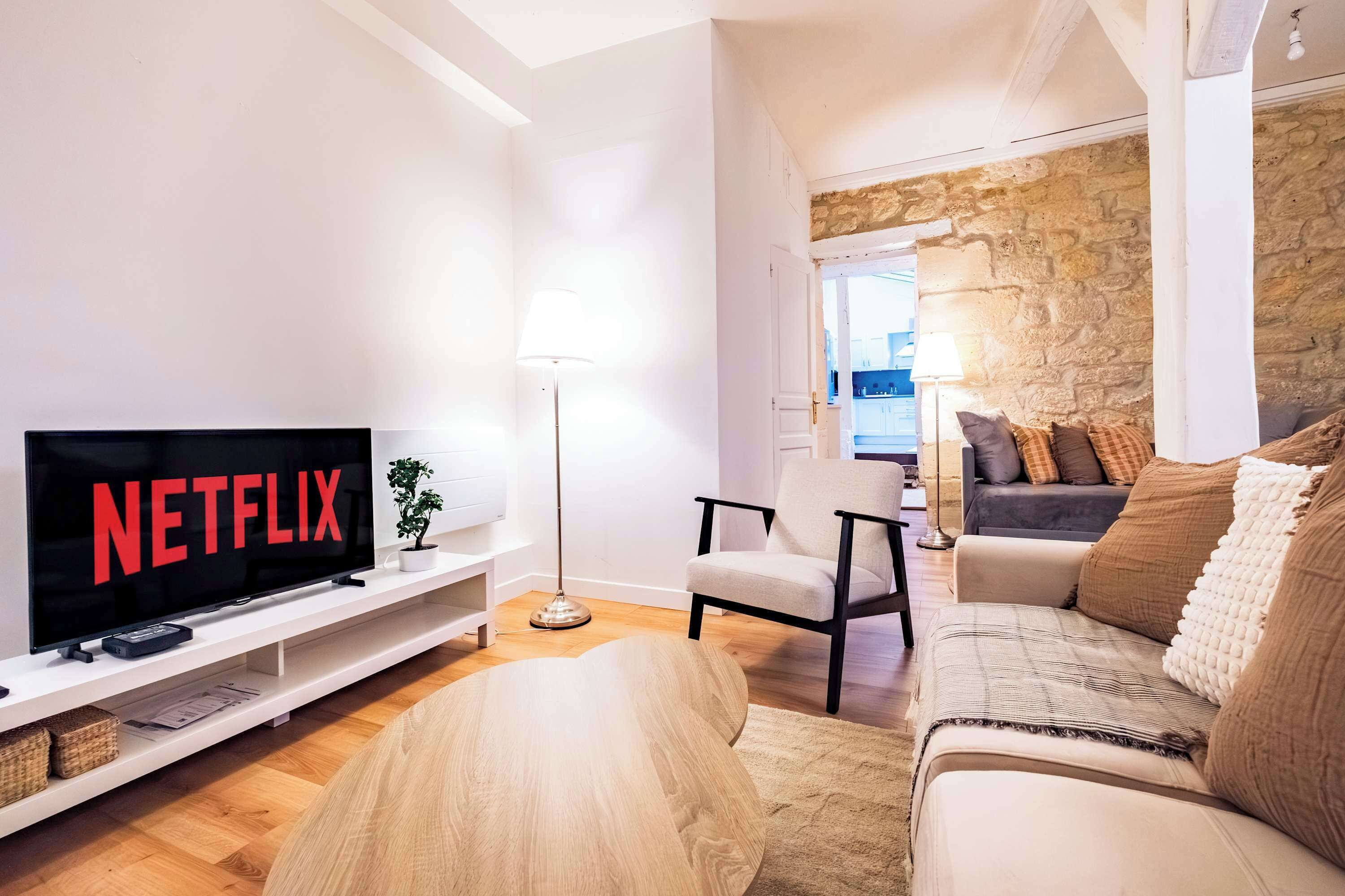 Ett modernt vardagsrum med en platt-TV som visar "NETFLIX", en vit soffa, ett soffbord i trä och en vägg av sten i bakgrunden.